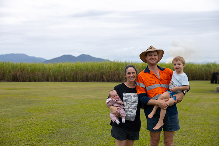 A young family of four stands in their backyard surrounded by sugar cane with mountains in the distance.