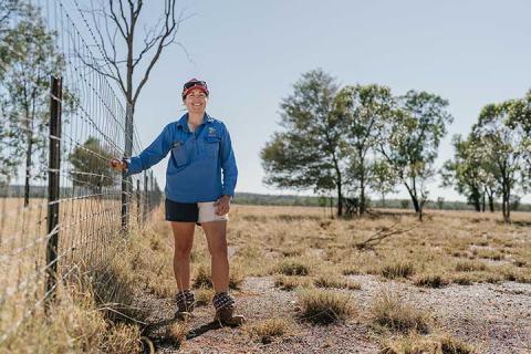 A young woman stands in a paddock next to a fence with her arm resting on the fence wire.